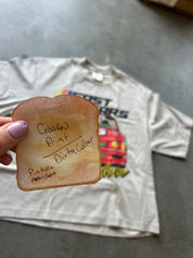 A hand with lavender nail polish holds a toasted bread slice-shaped marker with handwritten text, in front of a faded graphic t-shirt on a gray concrete floor.