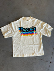 An off-white short-sleeve t-shirt laid flat on a gray concrete floor, featuring a retro-style graphic of the word 'Teach' in black with wavy rainbow stripes.