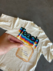 A hand holds a small printed toast graphic in front of a cream-colored t-shirt laid flat on a gray concrete floor. The t-shirt features a colorful retro-style rainbow graphic spelling 'Teach' in black.