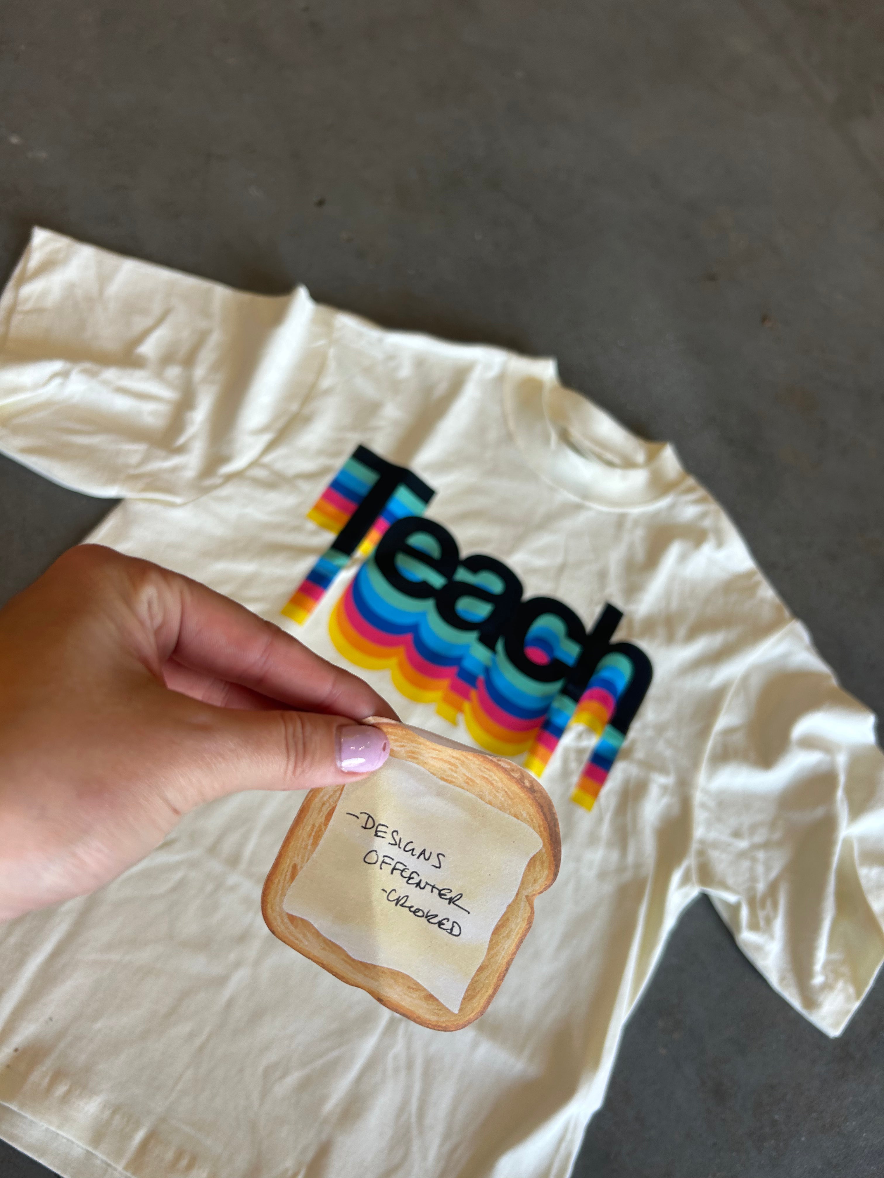 A hand holds a small printed toast graphic in front of a cream-colored t-shirt laid flat on a gray concrete floor. The t-shirt features a colorful retro-style rainbow graphic spelling 'Teach' in black.