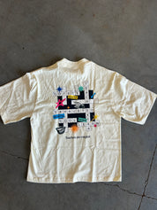 An off-white short-sleeve t-shirt with a colorful crossword puzzle graphic on the back, laid flat on a gray concrete floor.