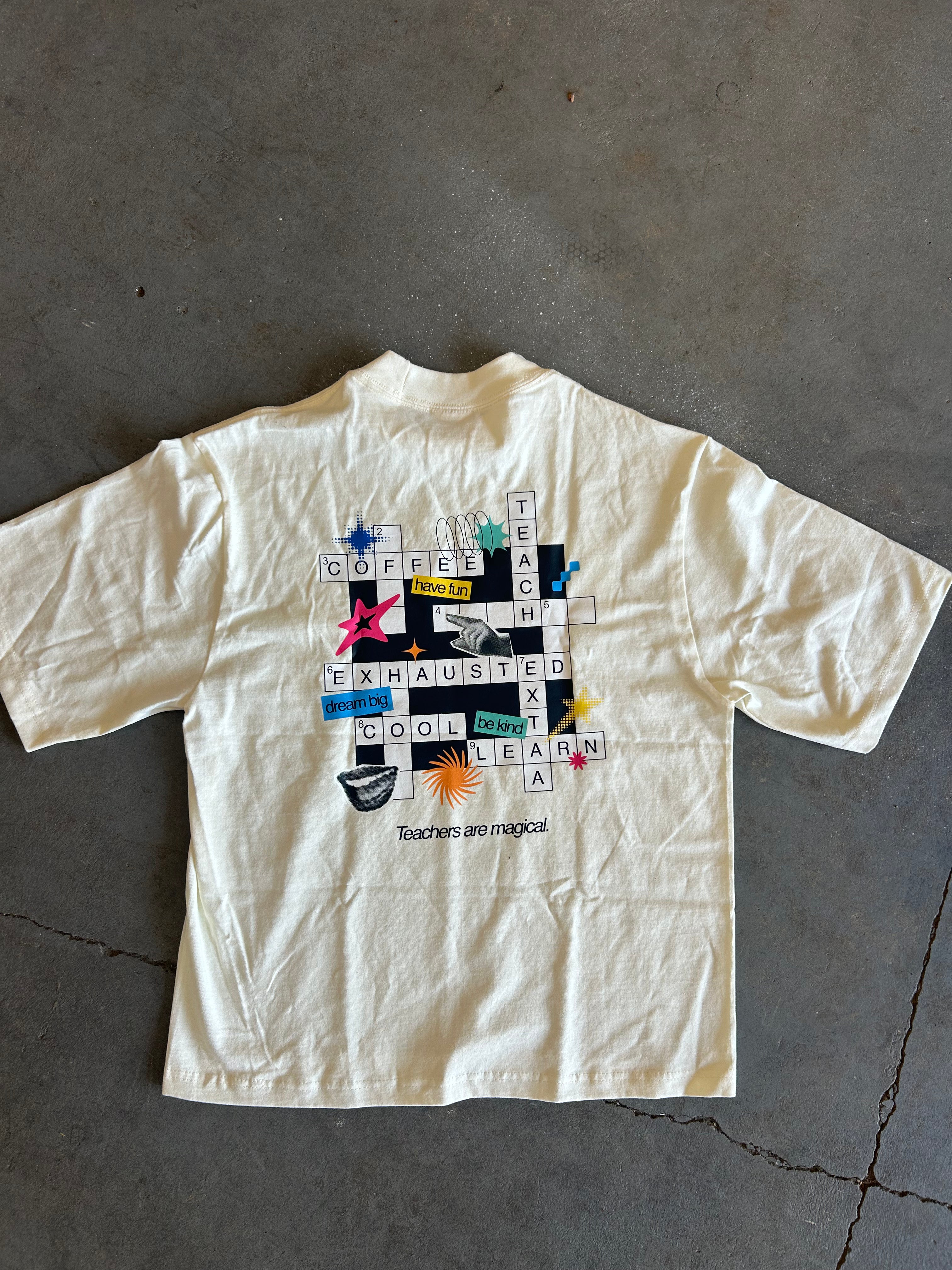 An off-white short-sleeve t-shirt with a colorful crossword puzzle graphic on the back, laid flat on a gray concrete floor.