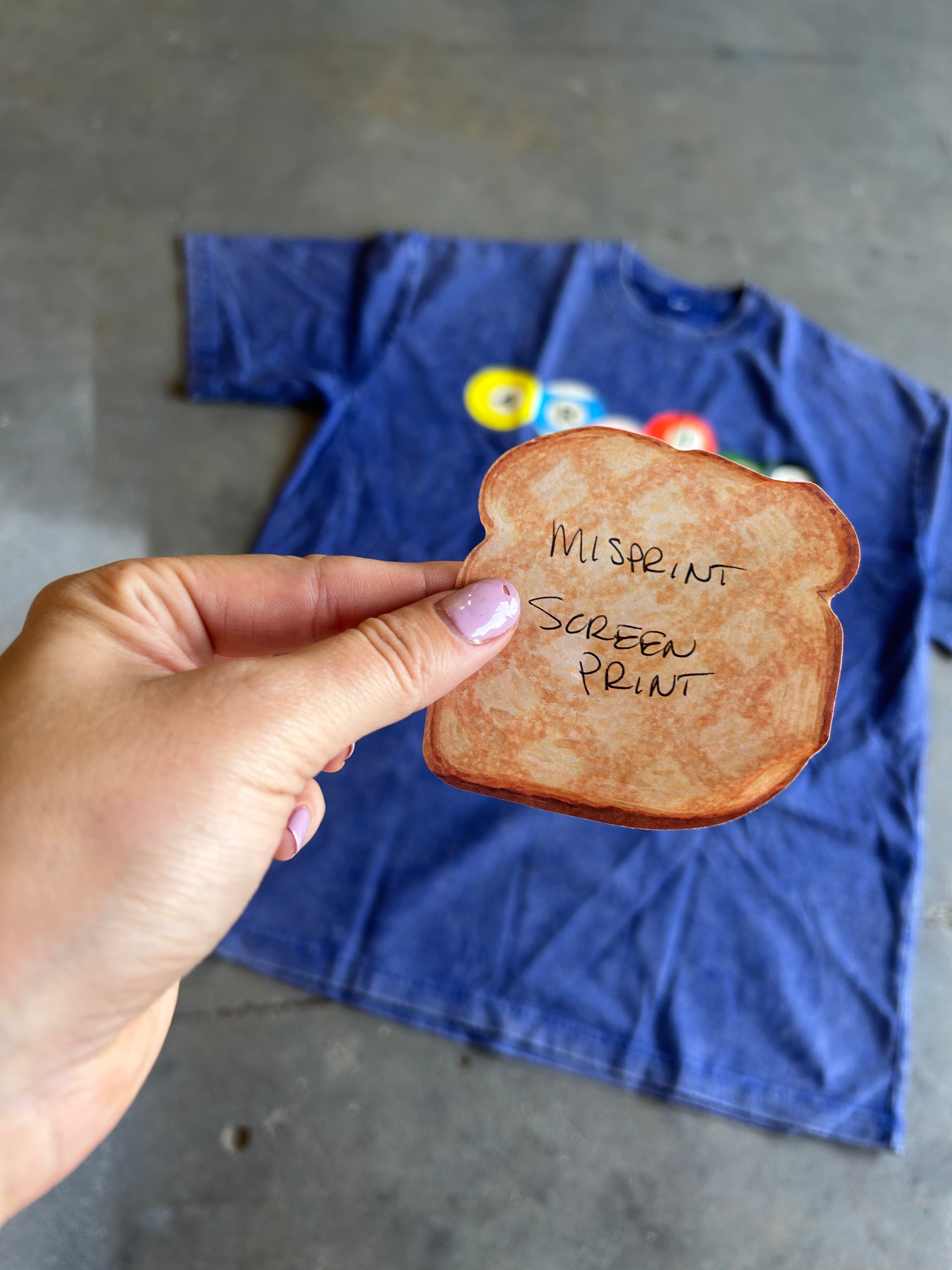 A hand holds a toasted bread slice prop in front of a blue t-shirt on a gray concrete floor. The bread has handwritten text reading 'MISSPRINT SCREEN PRINT'.