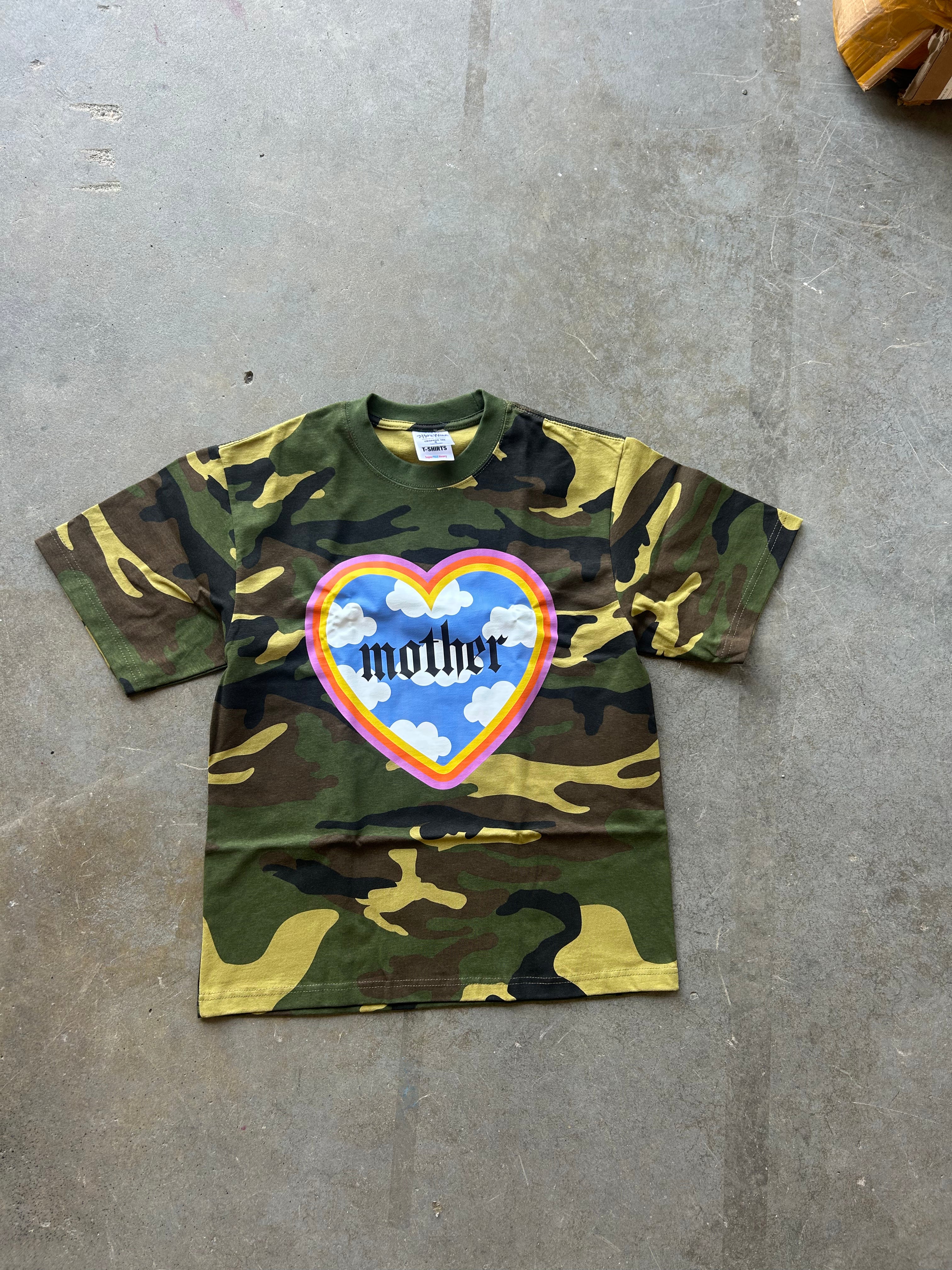 A short-sleeve camouflage t-shirt laid flat on a gray concrete floor. The shirt features a green, brown, and tan camo pattern with a large heart graphic on the chest containing the word 'mother' in black text over a blue sky and white clouds design.