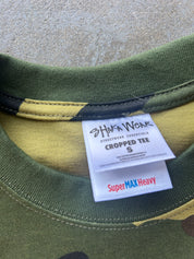 Close-up of the inside collar of a cropped t-shirt showing a white brand label that reads 'HAKA WEAR' and 'Super MAX Heavy'. The shirt features an olive green and yellow camouflage pattern.
