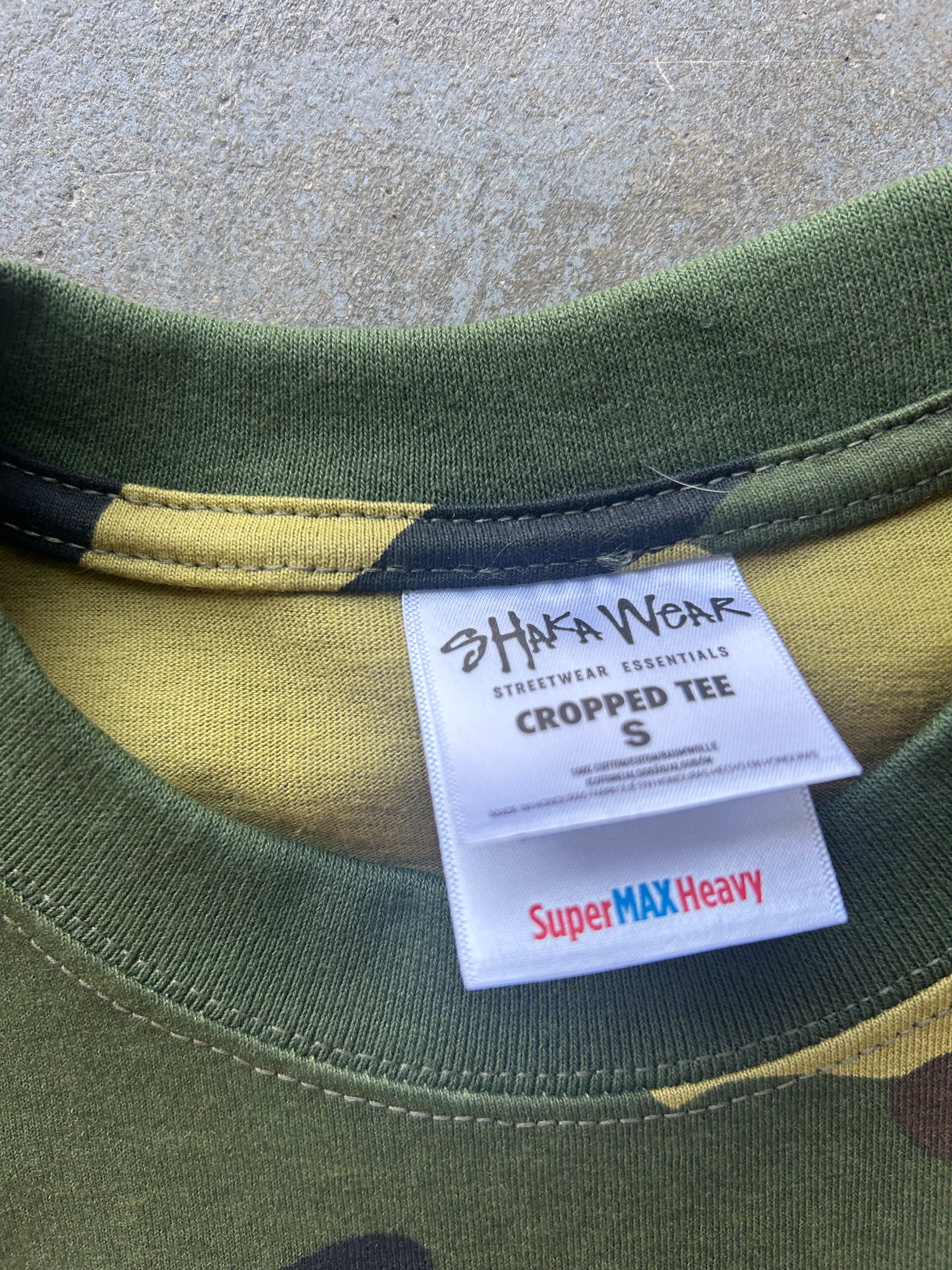 Close-up of the inside collar of a cropped t-shirt showing a white brand label that reads 'HAKA WEAR' and 'Super MAX Heavy'. The shirt features an olive green and yellow camouflage pattern.