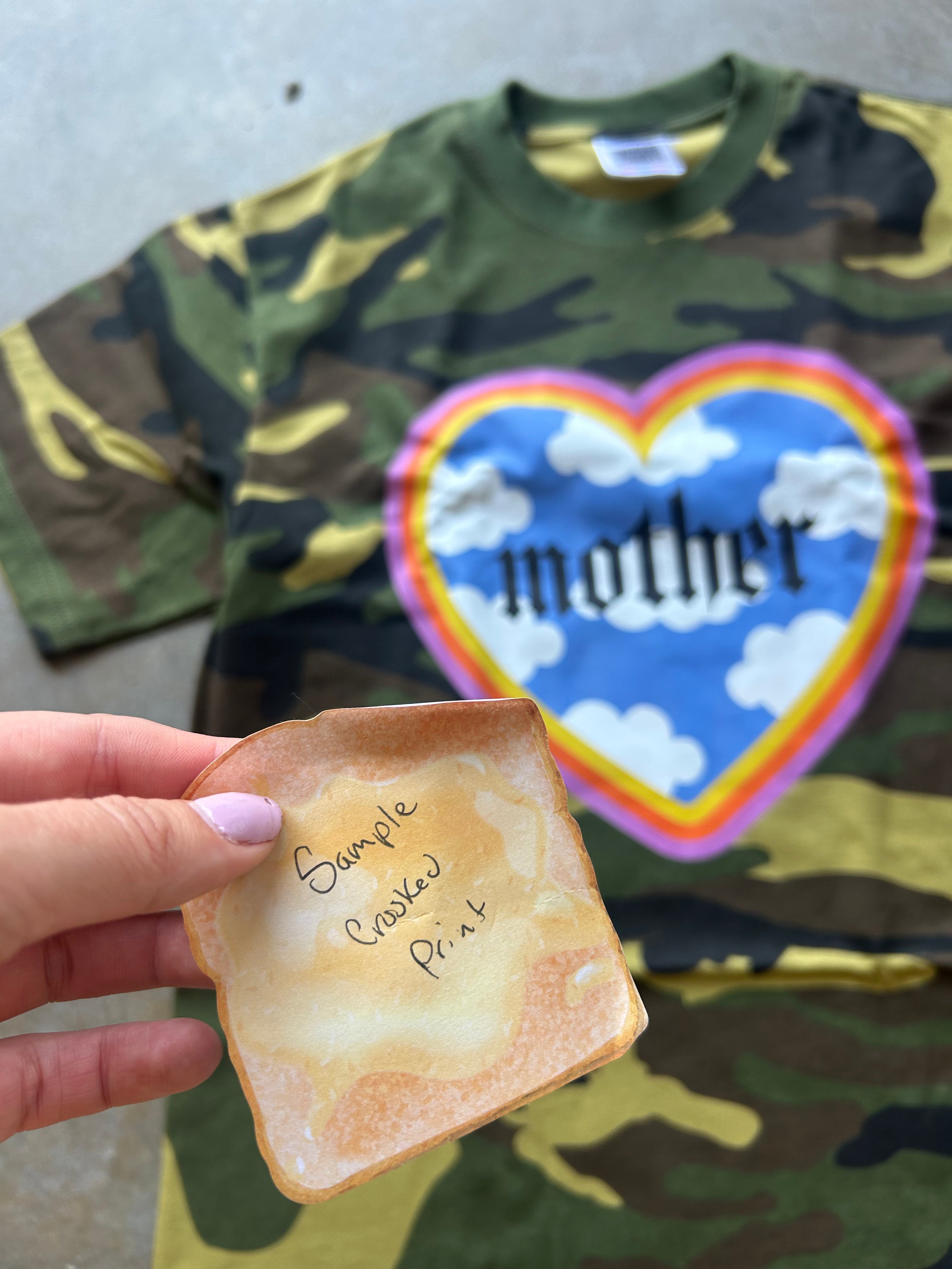 A camouflage T-shirt with a large heart graphic reading 'mother' is laid out on a surface, while a hand holds a toasted bread slice in front of it. The shirt has a green, brown, and tan camo pattern with a blue, pink, and yellow heart design.