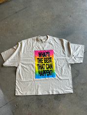 A cream-colored short-sleeve t-shirt with a colorful rainbow graphic on the chest that reads 'WHAT'S THE BEST THAT CAN HAPPEN?'. The shirt is laid flat on a gray concrete floor.
