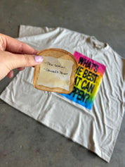 A hand with pink nail polish holds a printed toast-shaped sticker above a white t-shirt on a gray concrete floor. The sticker features a golden-brown toasted bread design with a cream center containing the handwritten quote '-PEN SOAM - ORCHIDED PRINT'.