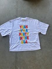 A white short-sleeve t-shirt with a colorful graphic text reading 'YEAH IT'S A PARTY IN THE USA' in red, blue, and yellow block letters, laid flat on a gray concrete floor.