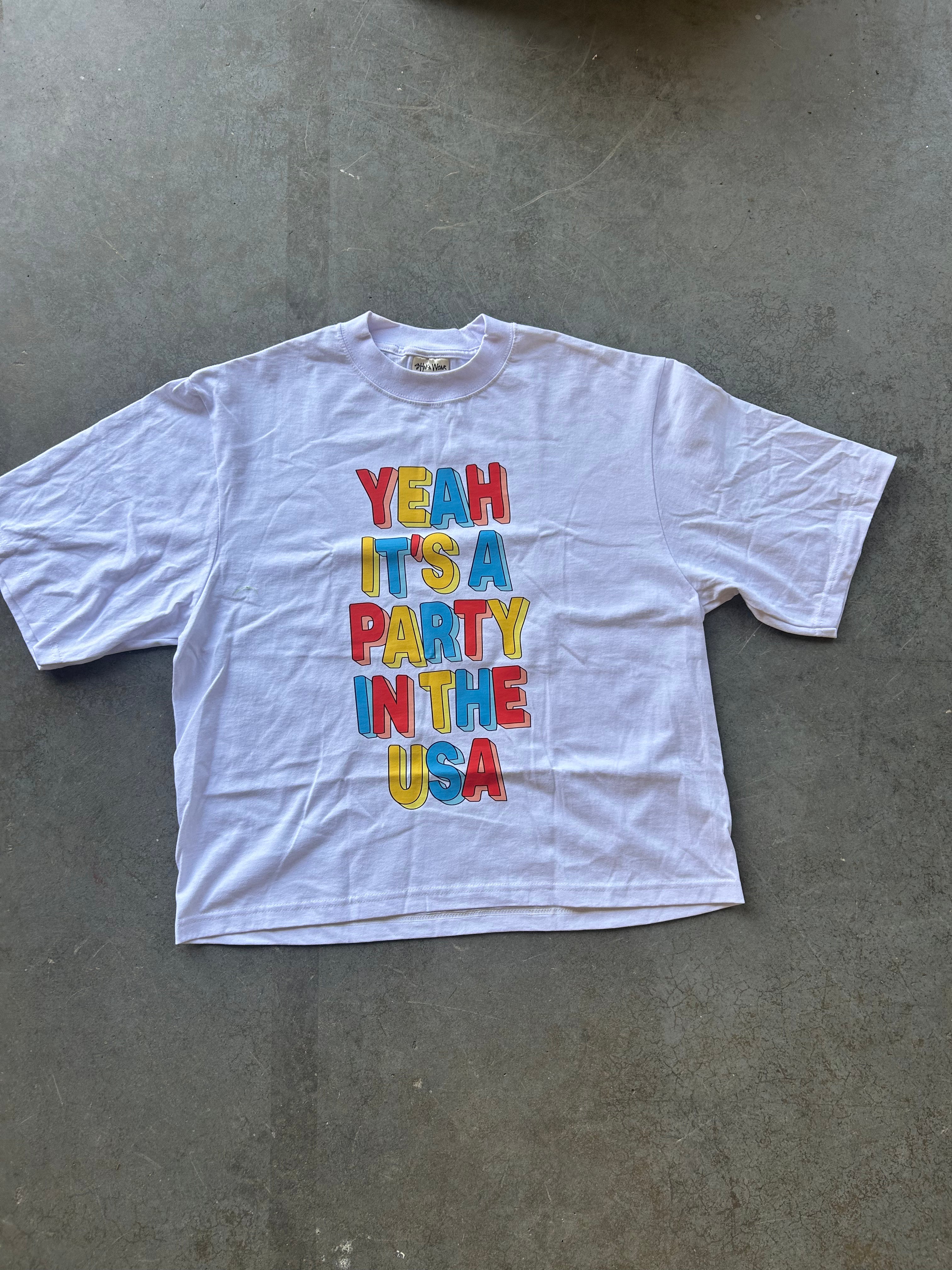 A white short-sleeve t-shirt with a colorful graphic text reading 'YEAH IT'S A PARTY IN THE USA' in red, blue, and yellow block letters, laid flat on a gray concrete floor.