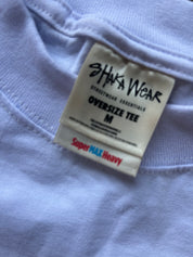 Close-up of a light blue oversized t-shirt tag reading 'SHAKA WEAR' and 'OVERSIZE TEE M' with 'Super MAX Heavy' printed in red and blue.