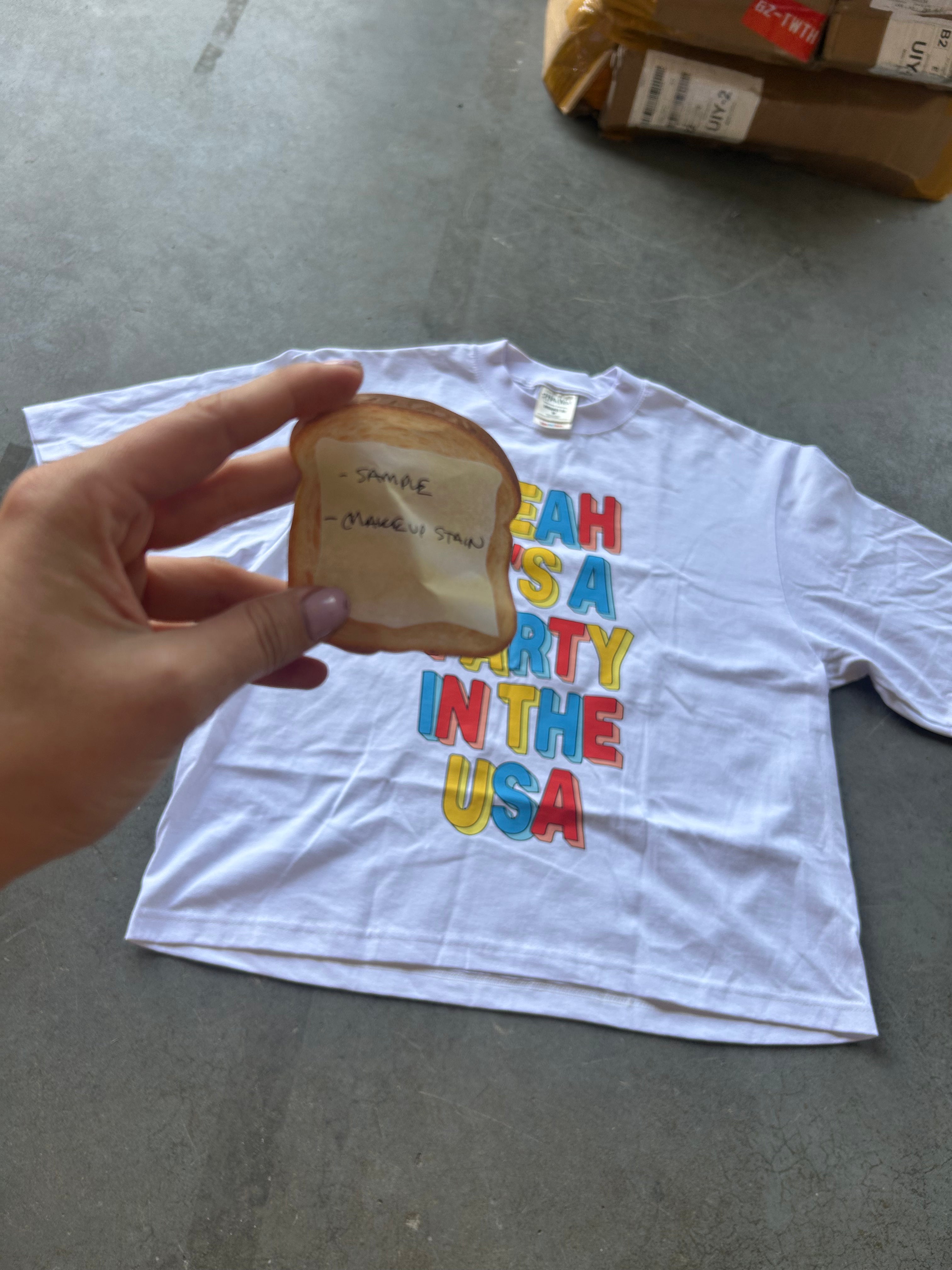 A white graphic t-shirt with a colorful 'MAKE AMERICA PARTY IN THE USA' print, held by a hand over a gray concrete floor.