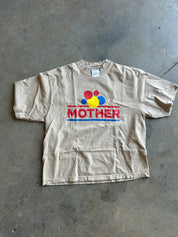 A beige short-sleeve t-shirt with a colorful graphic on the chest featuring the word 'MOTHER' in red, blue, and yellow, laid flat on a gray concrete floor.