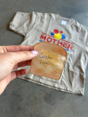 A hand holds a toasted bread slice with the handwritten text 'Crooked Logo' in front of a beige t-shirt featuring a colorful 'MOTHER' graphic on a gray concrete floor.