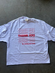 A white short-sleeve t-shirt with a red graphic print reading 'THANK YOU' repeated in outline and solid text, with 'for raising your hand' underneath, laid flat on a dark gray concrete surface.