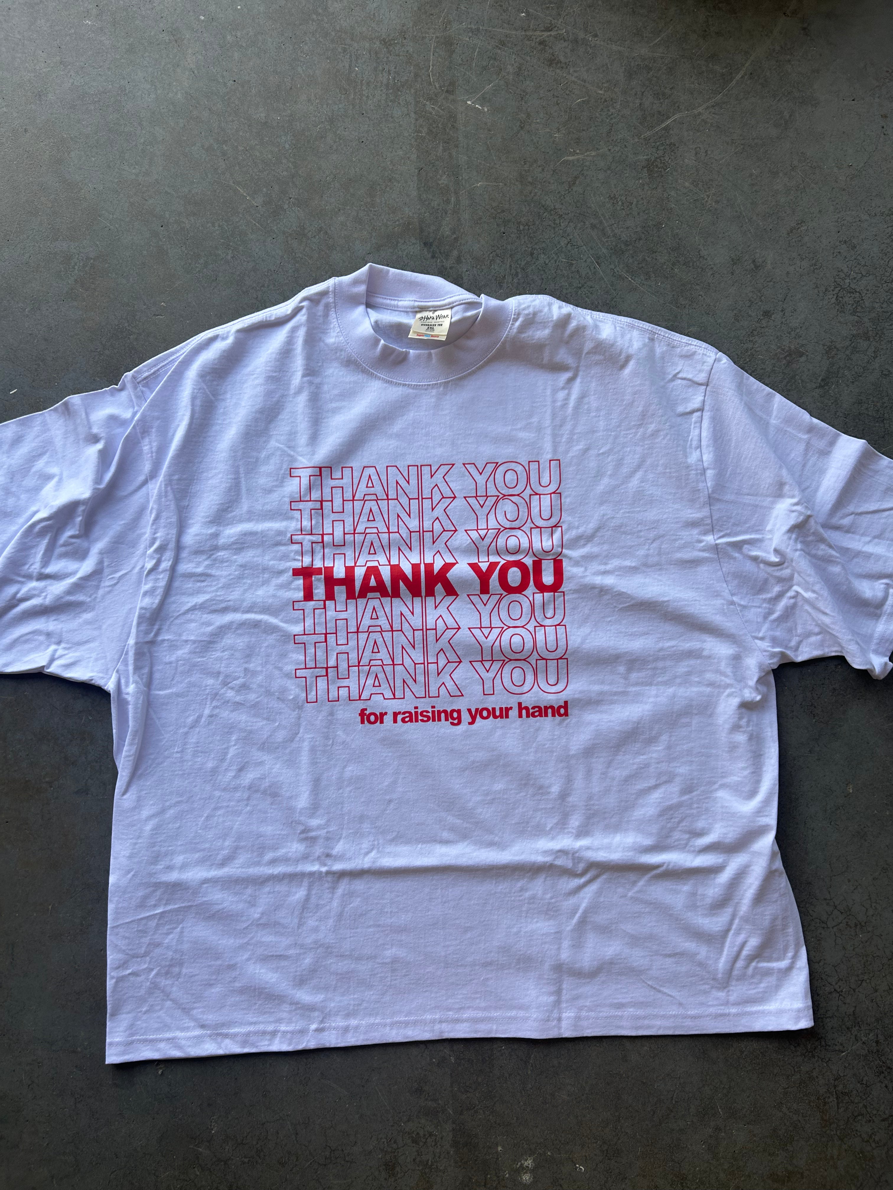 A white short-sleeve t-shirt with a red graphic print reading 'THANK YOU' repeated in outline and solid text, with 'for raising your hand' underneath, laid flat on a dark gray concrete surface.
