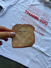 A hand holds a toasted bread slice with the word 'Cooked' written on it, in front of a white t-shirt printed with the phrase 'THANK YOU your hand' in red and pink text.