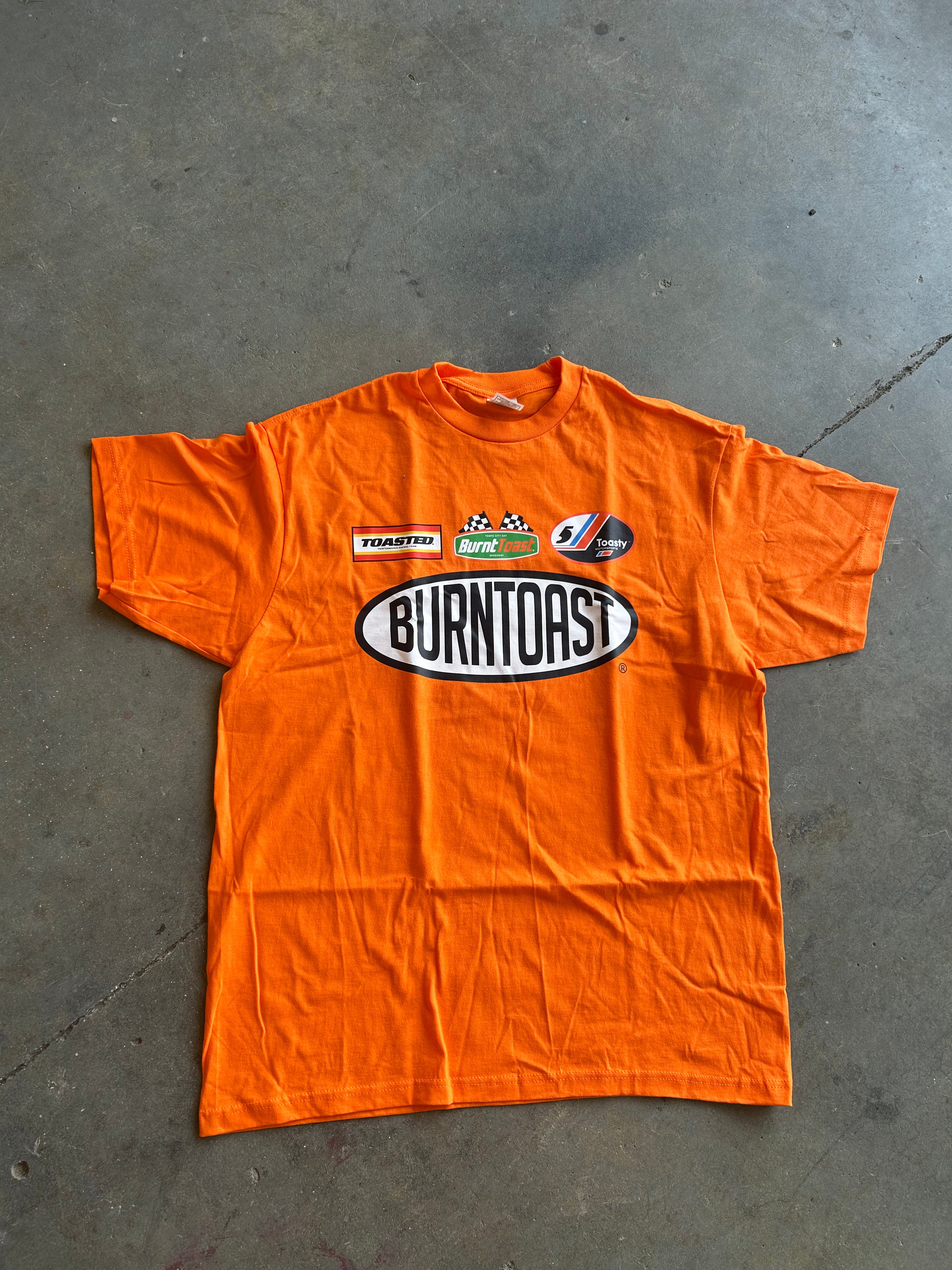 An orange short-sleeve t-shirt with a large 'Burntoast' oval logo and smaller sponsor logos on the chest, laid flat on a gray concrete floor.