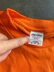 Close-up of an orange crewneck t-shirt showing the inner neck label for Johnnie Wear Active Soft Silk Cotton, with a finger pointing at the label.
