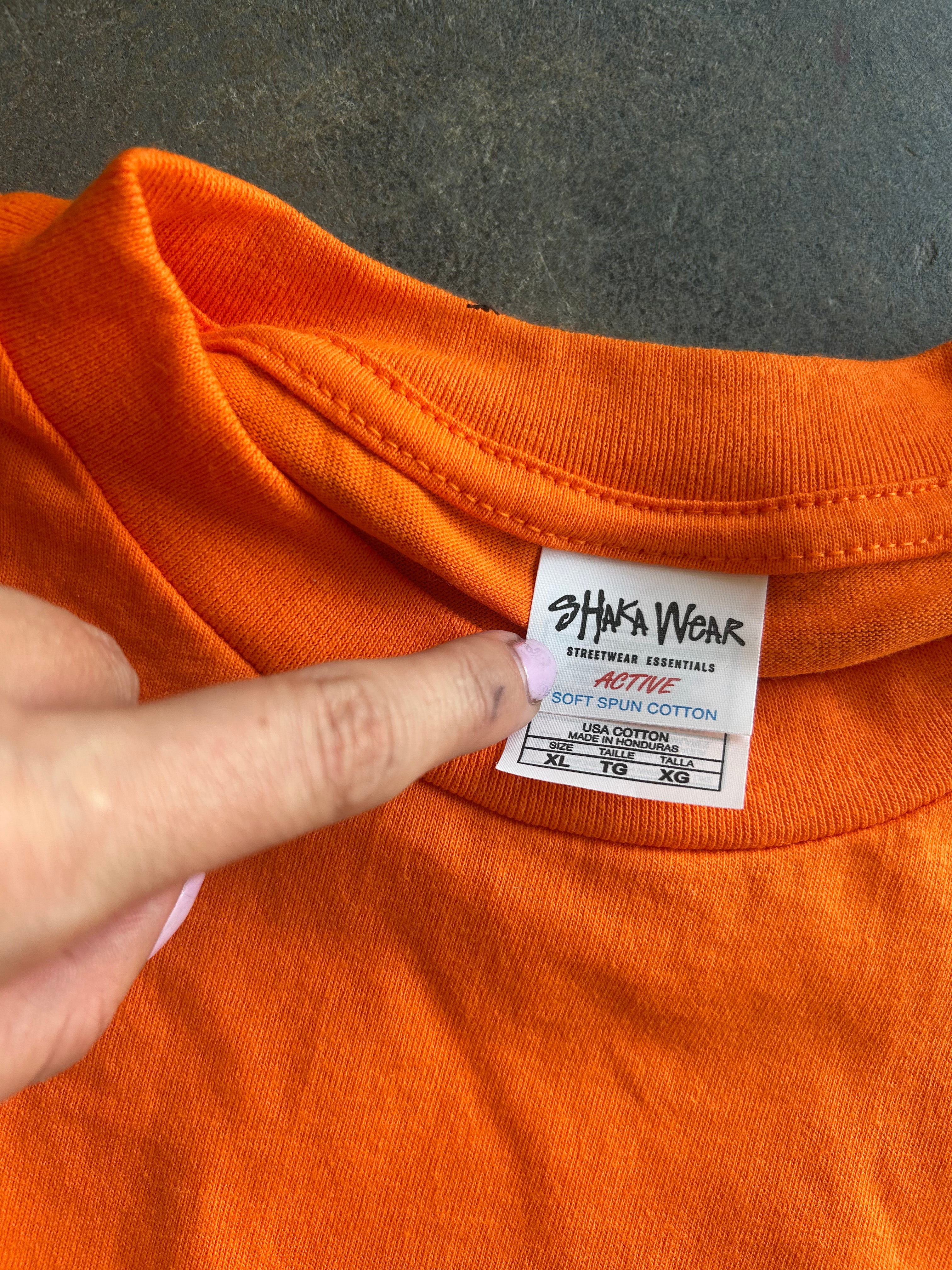 Close-up of an orange crewneck t-shirt showing the inner neck label for Johnnie Wear Active Soft Silk Cotton, with a finger pointing at the label.