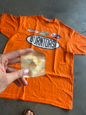 An orange graphic t-shirt with 'BURNTOAST' printed on the front is laid flat on a gray concrete floor. A hand is holding a small, handwritten note in a piece of parchment paper in the foreground.