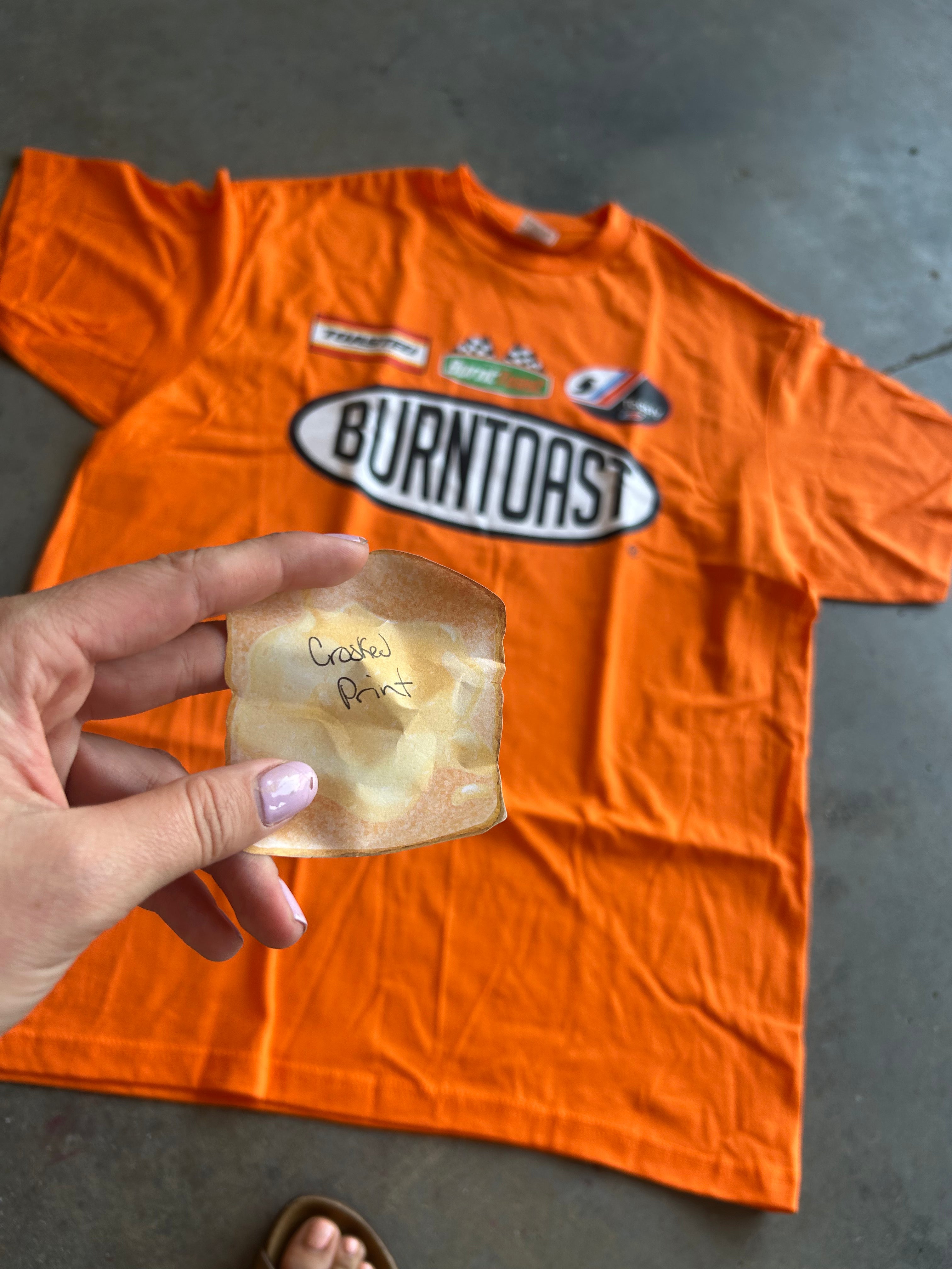 An orange graphic t-shirt with 'BURNTOAST' printed on the front is laid flat on a gray concrete floor. A hand is holding a small, handwritten note in a piece of parchment paper in the foreground.