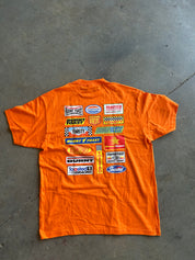 An orange short-sleeve T-shirt laid flat on a gray concrete floor, featuring a large collage of various racing logos and decals printed on the back.