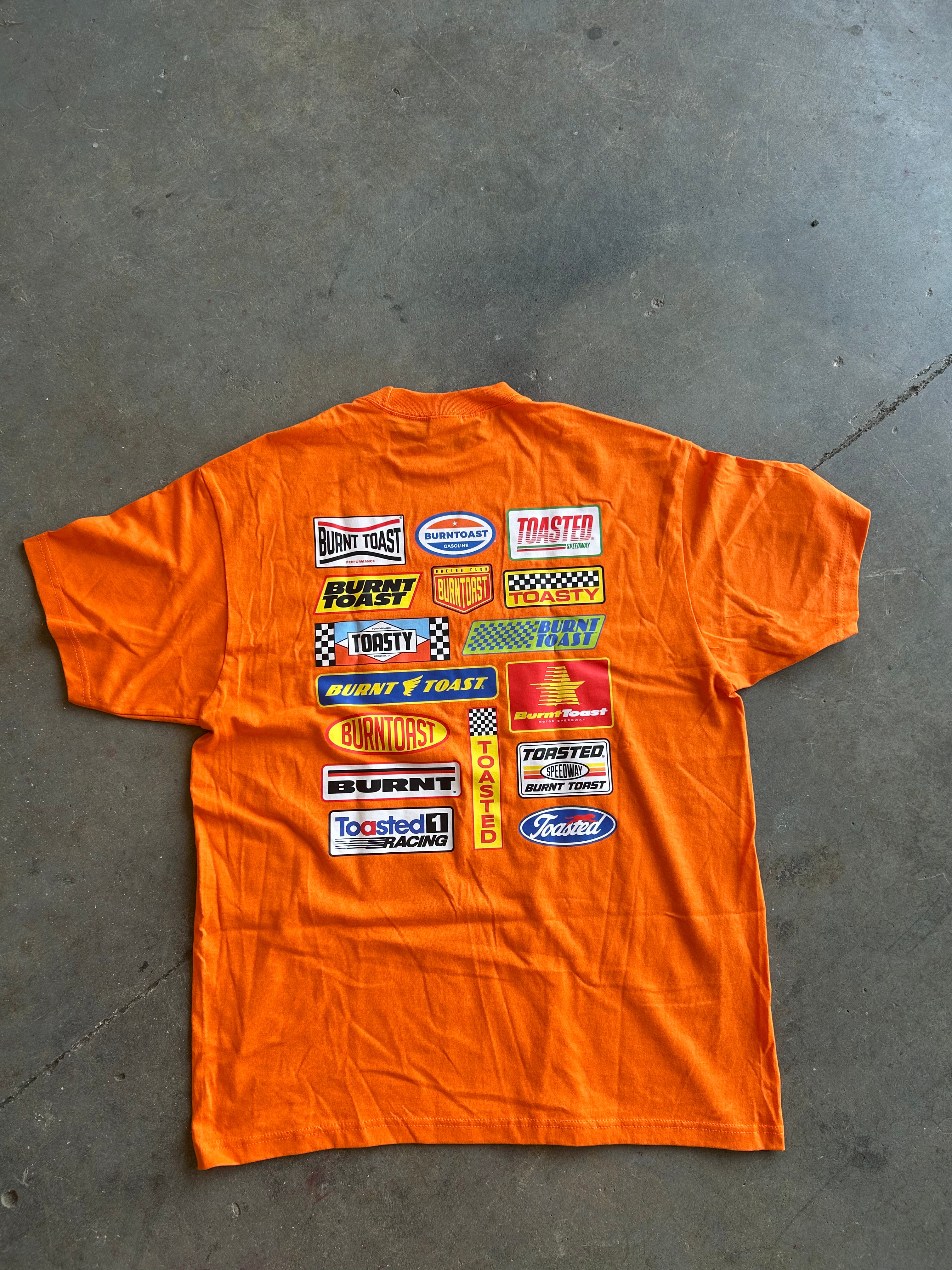 An orange short-sleeve T-shirt laid flat on a gray concrete floor, featuring a large collage of various racing logos and decals printed on the back.