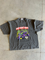 A dark gray graphic t-shirt laid flat on a concrete floor. The shirt features a graphic of a purple monster truck with green flames and the text 'DIRT DON'T HURT' in red and white.