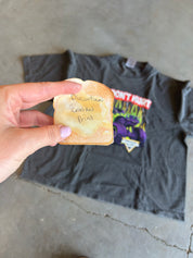 A hand holds a toasted bread slice with handwritten text over a dark gray graphic t-shirt laid on a concrete floor.