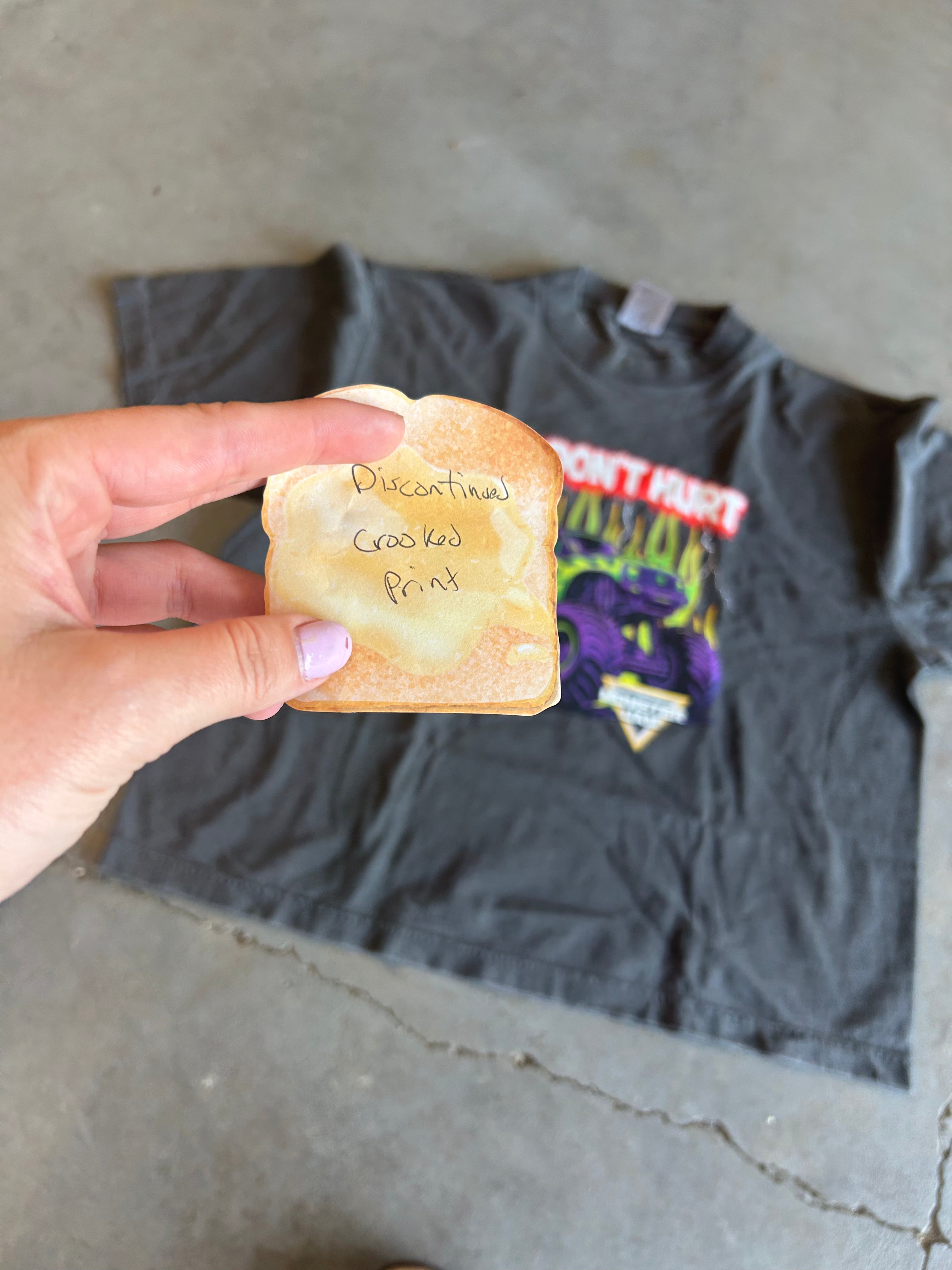 A hand holds a toasted bread slice with handwritten text over a dark gray graphic t-shirt laid on a concrete floor.