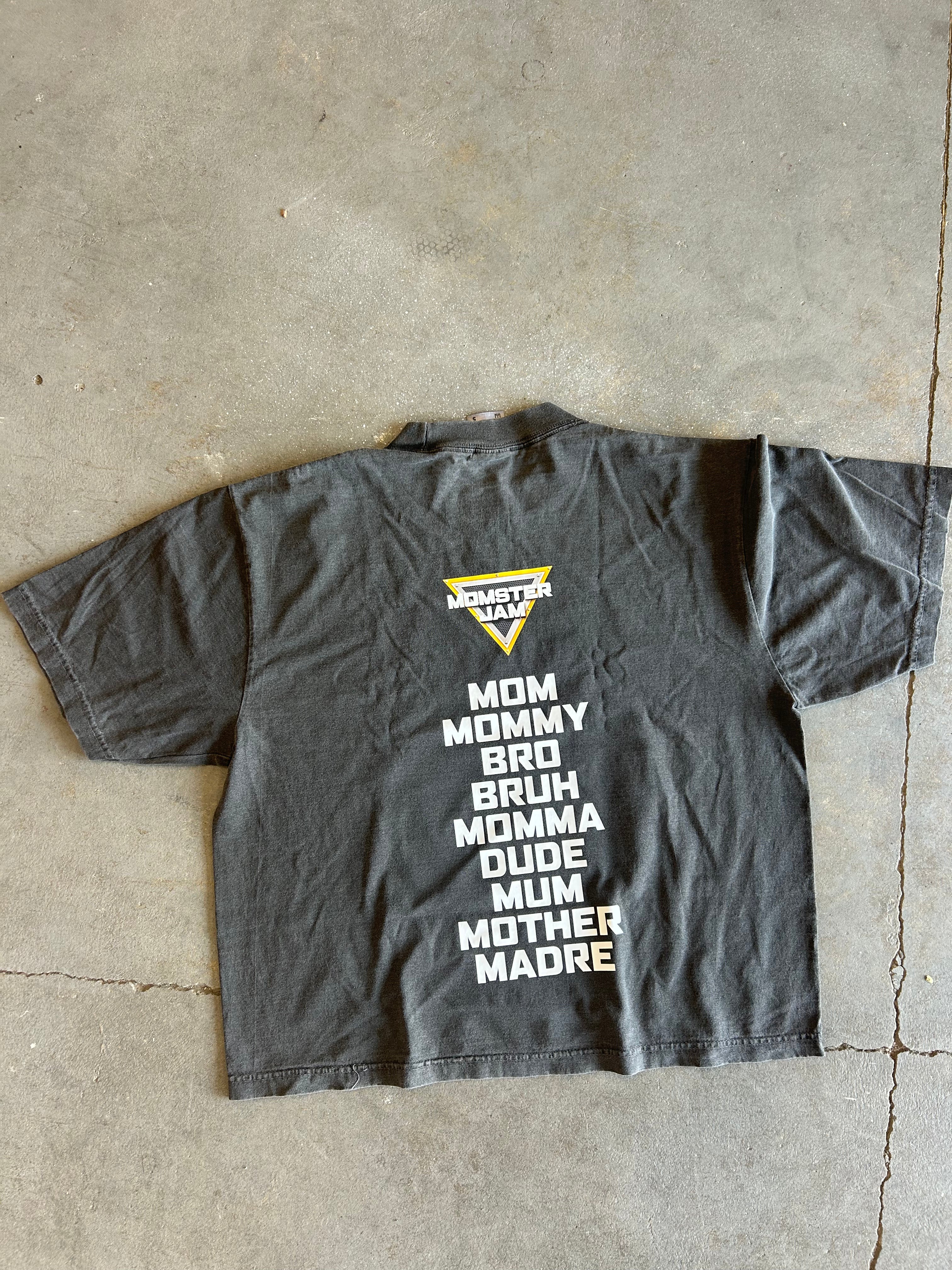 A dark gray t-shirt laid flat on a concrete floor, showing the back side with a yellow and white Monster Mayhem logo and a list of humorous roles in white text.