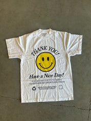 A white short-sleeve graphic T-shirt laid flat on a gray concrete floor. The shirt features a large yellow smiley face in the center with the words 'THANK YOU!' above it and 'Have a Nice Day!' below in black text.