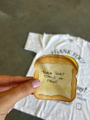 A hand holds a novelty sandwich toy shaped like a slice of toast with a printed yellow sticky note that reads, 'BLACK SHIRT STAINS FRONT.' In the blurred background, a white graphic t-shirt is laid on a concrete floor.