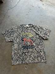 A vintage short-sleeve graphic t-shirt with a snakeskin pattern, laid flat on a concrete floor. The shirt features a yellow and red graphic on the chest with text that reads 'Running Wild' and 'MONEY IS BUSINESS'.