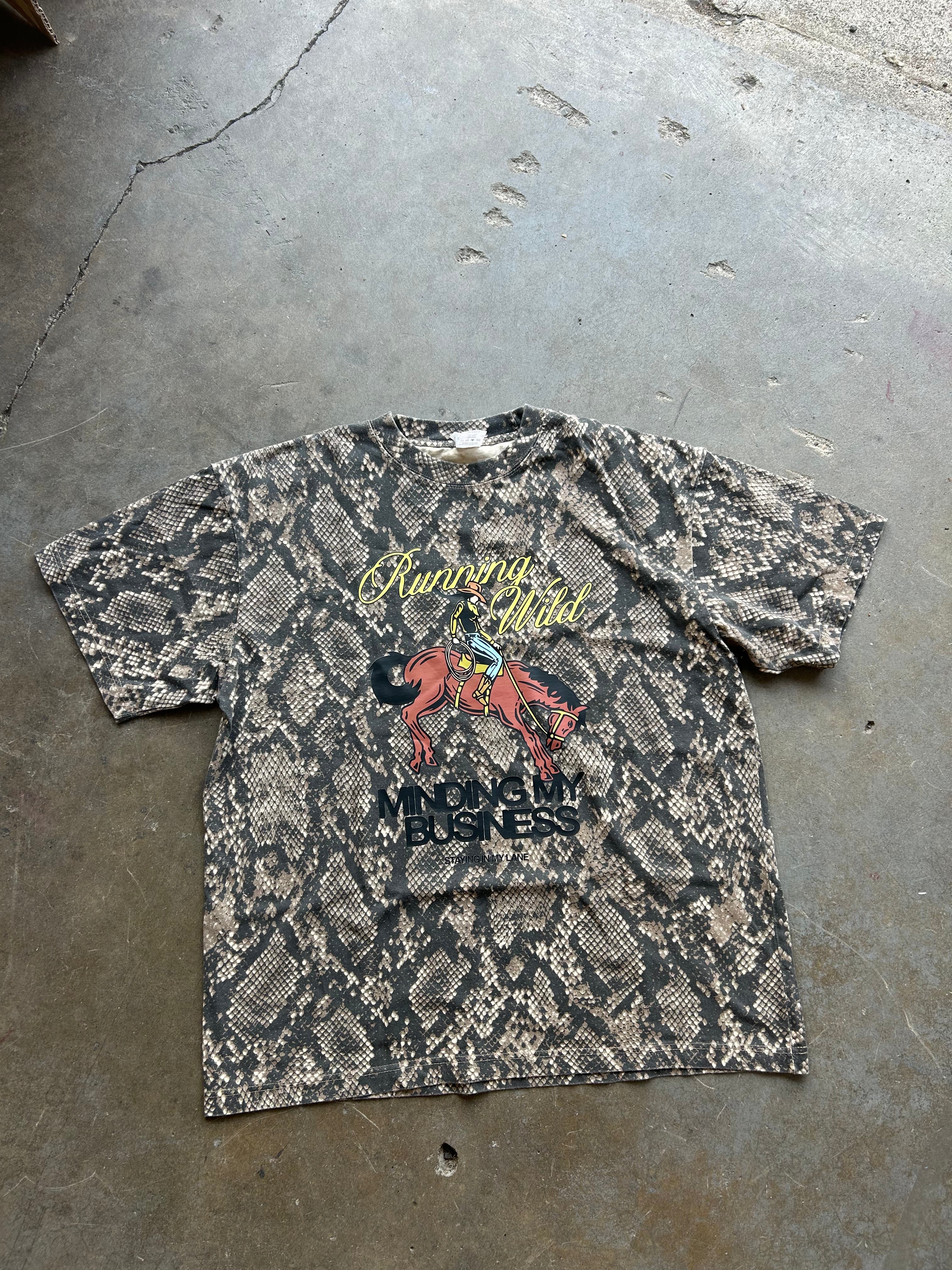 A vintage short-sleeve graphic t-shirt with a snakeskin pattern, laid flat on a concrete floor. The shirt features a yellow and red graphic on the chest with text that reads 'Running Wild' and 'MONEY IS BUSINESS'.