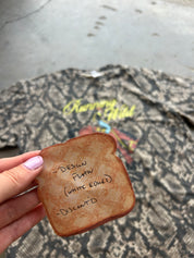 A hand with light purple nail polish holds a slice of toasted bread-shaped item in front of a snakeskin-patterned sweatshirt with yellow text. The item has handwritten text on it reading '- DESIGN PLAN (WHITE EDGES)' and '- DISCART'.