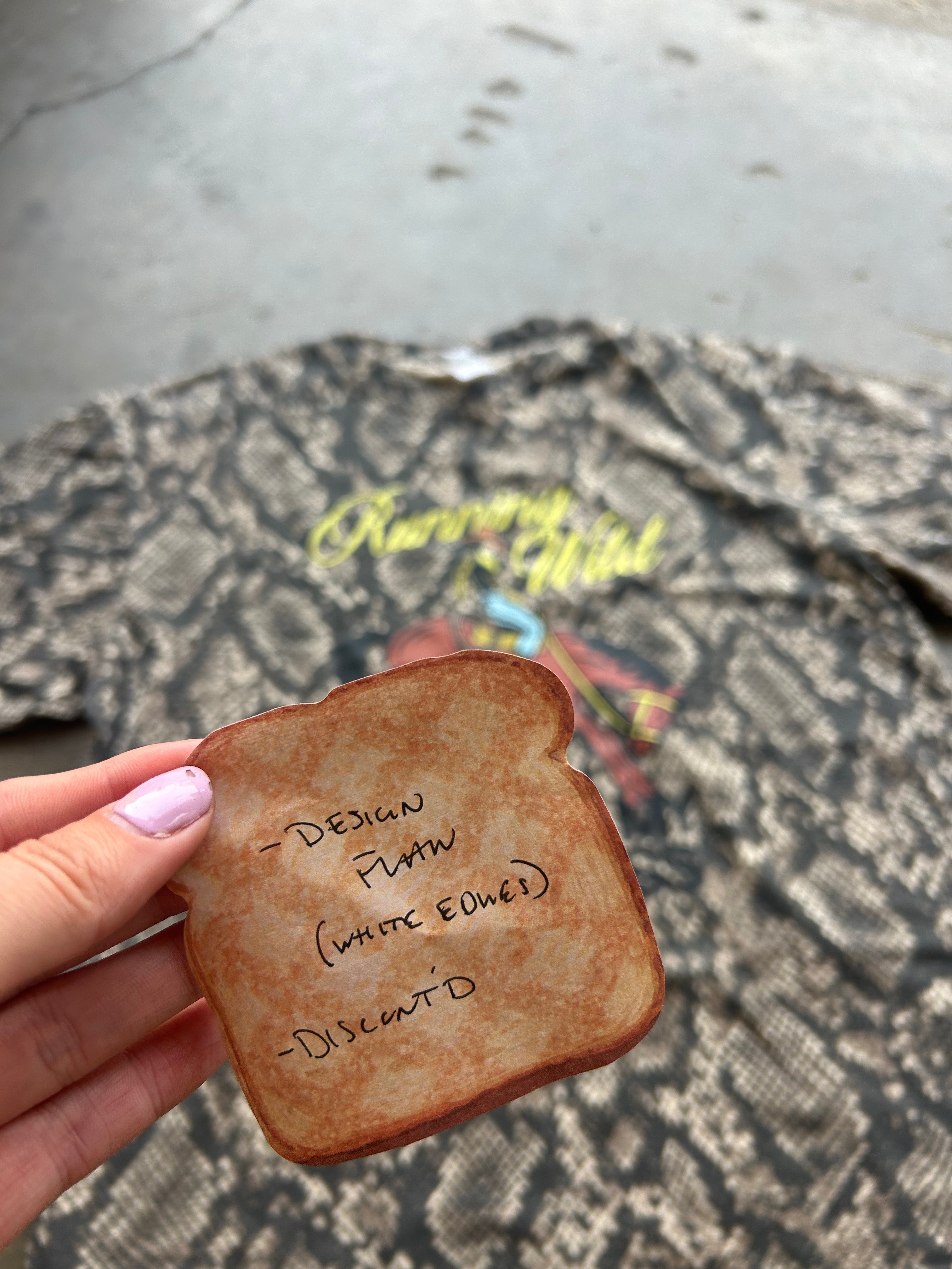 A hand with light purple nail polish holds a slice of toasted bread-shaped item in front of a snakeskin-patterned sweatshirt with yellow text. The item has handwritten text on it reading '- DESIGN PLAN (WHITE EDGES)' and '- DISCART'.