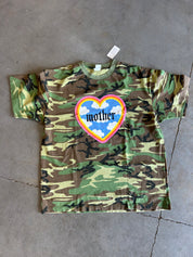 A short-sleeve camouflage t-shirt laid flat on a gray concrete floor. The shirt features a green, brown, and tan camo pattern with a central graphic of a heart containing blue clouds, a rainbow border, and the word 'mother' in black gothic font.