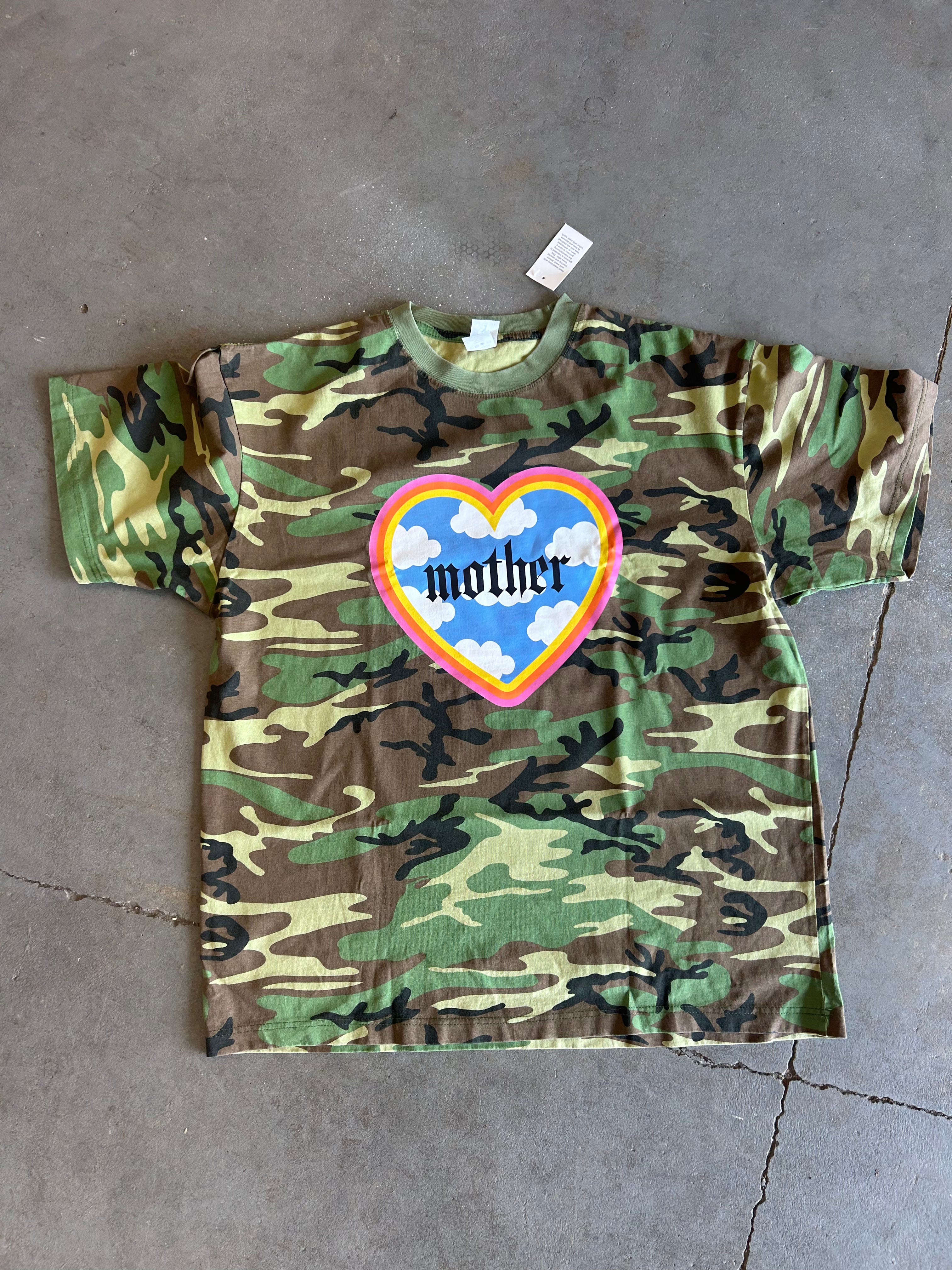 A short-sleeve camouflage t-shirt laid flat on a gray concrete floor. The shirt features a green, brown, and tan camo pattern with a central graphic of a heart containing blue clouds, a rainbow border, and the word 'mother' in black gothic font.