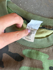 A close-up of a camouflage crewneck shirt being held at the collar by a hand, showing the inner care label which reads XLARGE.