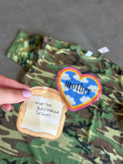 A hand holds a toasted bread slice mockup with a handwritten note over a camouflage t-shirt. The shirt features a colorful heart patch with the word 'mother' and a blue graphic.