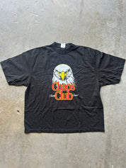 A black short-sleeve graphic t-shirt laid flat on a gray concrete floor. The shirt features a graphic of a bald eagle's head above the text 'Chaos Club' in orange-red and white.