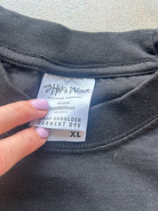 Close-up of a black garment dye label by Shaka Ngak attached to a black t-shirt collar, held by a hand with light purple nail polish against a concrete background.