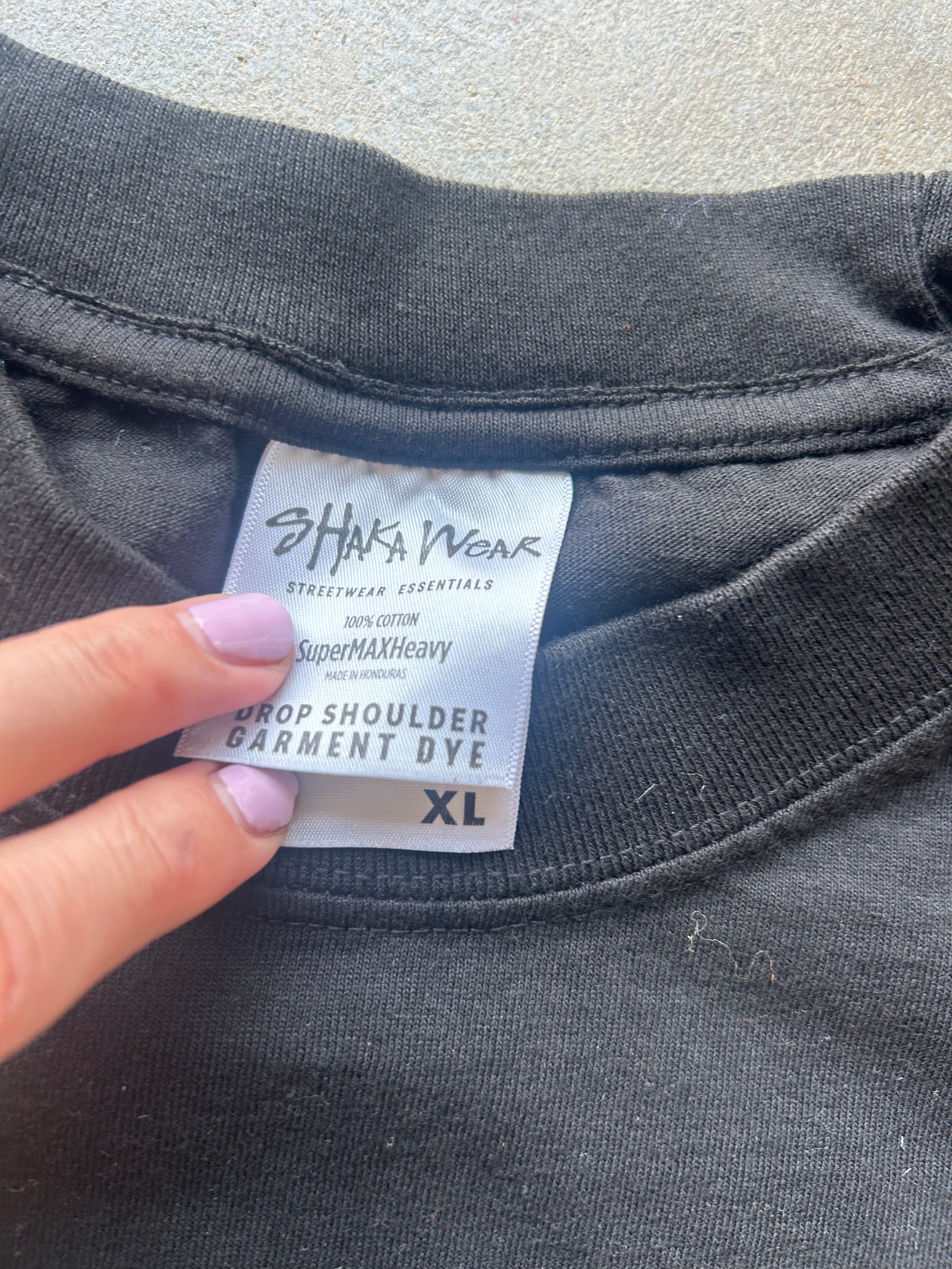 Close-up of a black garment dye label by Shaka Ngak attached to a black t-shirt collar, held by a hand with light purple nail polish against a concrete background.