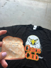 A hand holds a small novelty toast-shaped item with handwritten text, while a black graphic T-shirt with an eagle and the words 'Chaos Club' lies blurred in the background on a concrete surface.