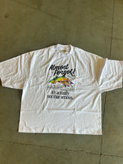 A white short-sleeve graphic t-shirt laid flat on a concrete floor. The shirt features a colorful rainbow fish illustration and the text 'Almost Forgot! it's actually not that serious.