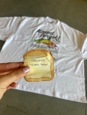 A hand holds a toasted sandwich bread slice with a yellow sticky note that reads 'COOKED - BLACK STARS' in front of a white graphic t-shirt on a concrete floor.