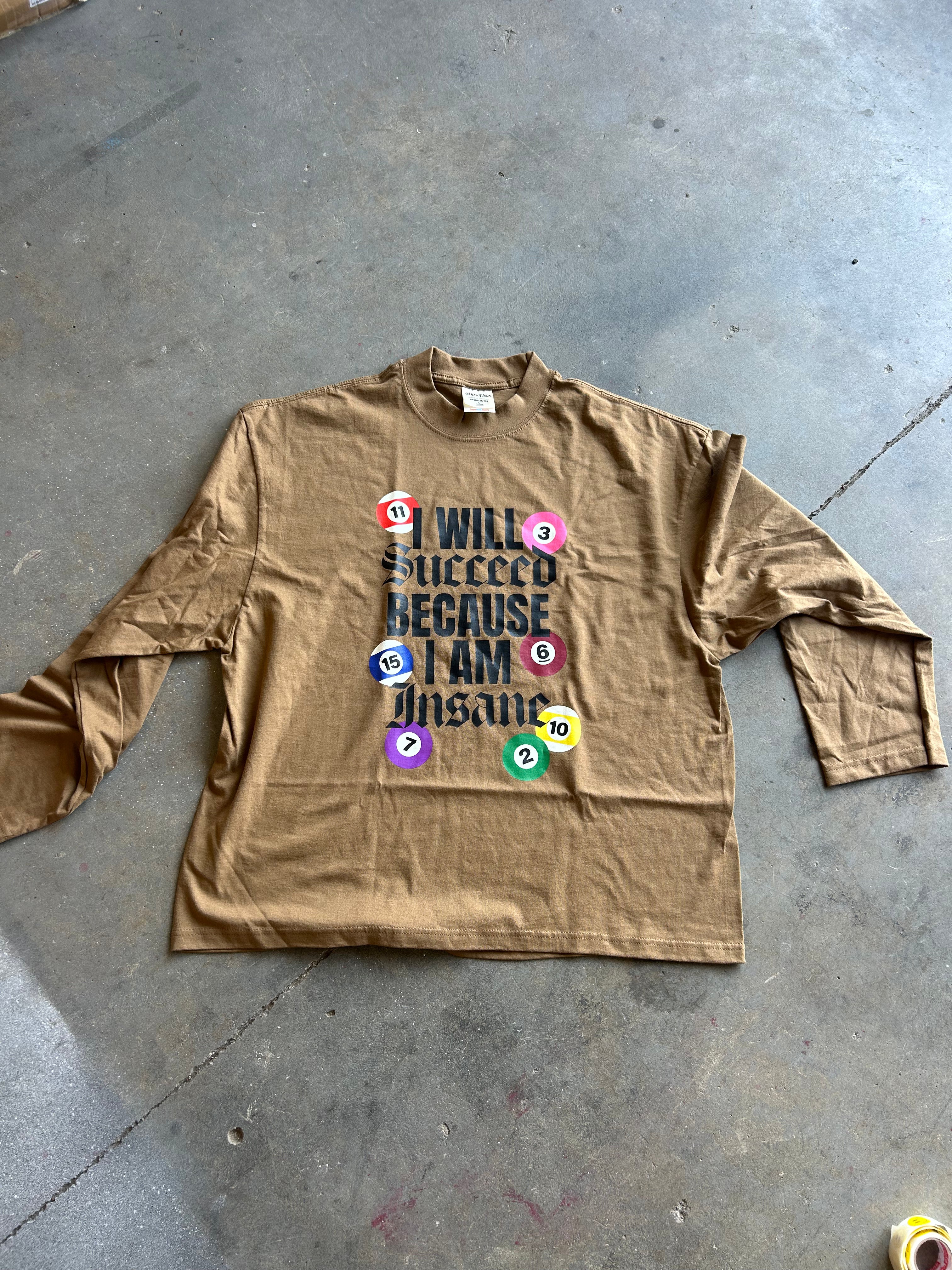 A tan long-sleeve graphic t-shirt laid flat on a gray concrete floor. The shirt features a graphic print with the text 'I WILL SUCCEED BECAUSE I AM a dinosaur' in black, surrounded by colorful billiard balls numbered 1 through 8.